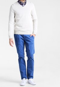 Personne portant un pull blanc à col en V, un pantalon bleu, des baskets blanches et une chemise à carreaux, se tenant avec une main dans la poche.