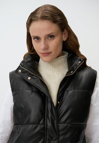 Gilet matelassé noir avec un col haut, doté d'une fermeture éclair, de boutons pression dorés et d'une texture brillante. Porté par-dessus un pull crème à côtes.