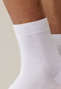 Zbliżenie na kostkę osoby, która ma na sobie gładką, białą skarpetkę sięgającą kostek z prążkowanym ściągaczem na neutralnym tle.