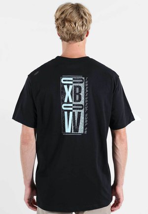 Personne blonde portant un T-shirt noir avec un logo stylisé "XBW" bleu et blanc et le texte "Independent Surf Co." au dos.