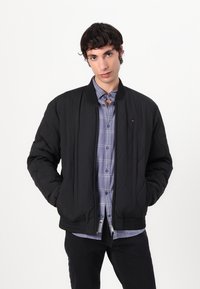 Black quilted bomber jacket with a ribbed collar and cuffs, featuring zippered pockets and subtle branding on the chest. Layered over a plaid shirt.