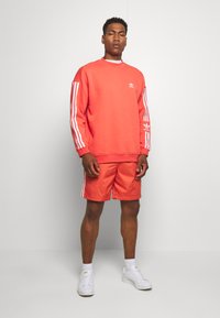 Felpa color corallo con logo adidas bianco e strisce bianche sulle maniche; pantaloni corti corallo coordinati. Modello che indossa sneakers bianche e calzini alla caviglia.