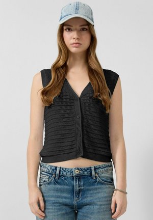 Młoda kobieta w jasnoniebieskiej dżinsowej czapce, czarnej, bezrękawnikowej, zapinanej na guziki kamizelce i niebieskich dżinsach na jednolitym tle.