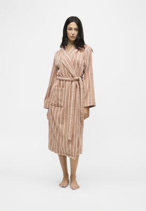 Femme debout pieds nus, portant un peignoir rose mi-genoux avec des rayures blanches verticales et une ceinture nouée, sur un fond blanc uni.