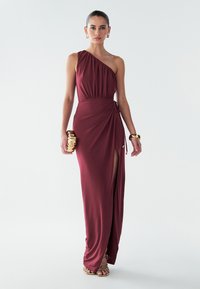 Bordové šaty s jedným ramienkom, s nazbieraným živôtikom, bočným rozparkom a detailom viazaného pása. Modelka drží zlaté psaníčko. Hladká látka s elegantným dizajnom.