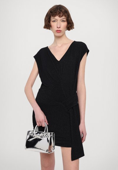 Robe noire courte avec un décolleté en V et un design drapé, présentant un détail en nœud à la taille. Le mannequin tient un petit sac à main argenté et réfléchissant.