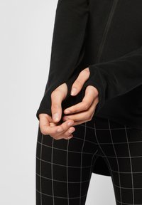 Mains ajustant les trous pour les pouces sur un haut noir à manches longues associé à des leggings noirs avec un motif à carreaux blanc.