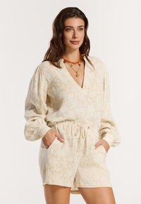 Salopette di lino beige con scollatura a V, maniche a palloncino e vita elastica. Presenta una texture floreale e tasche laterali. Accentuata con collane a strati.