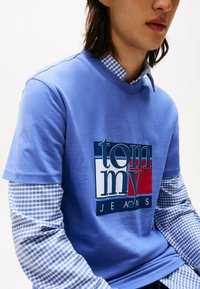 Hombre vistiendo una camiseta de Tommy Jeans azul y roja, superpuesta sobre una camisa de manga larga de cuadros en azul y blanco, mostrado desde el pecho hasta el cuello.