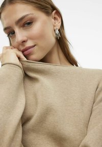 Maglione lavorato a maglia beige con scollatura ampia, tessuto morbido e vestibilità aderente. La modella tiene leggermente il colletto, rivelando degli orecchini a cerchio lucidi.