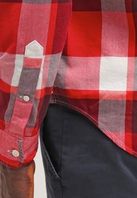 Manche de chemise à carreaux rouge, blanc et gris avec poignet boutonné à côté de la poche d’un pantalon bleu marine sur le bras et la taille d’une personne.