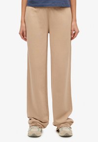 Beżowe szerokie spodnie dresowe o miękkiej fakturze i elastycznym pasku w talii, noszone ze szarymi tenisówkami. Gładki materiał bez widocznych wzorów ani zdobień.