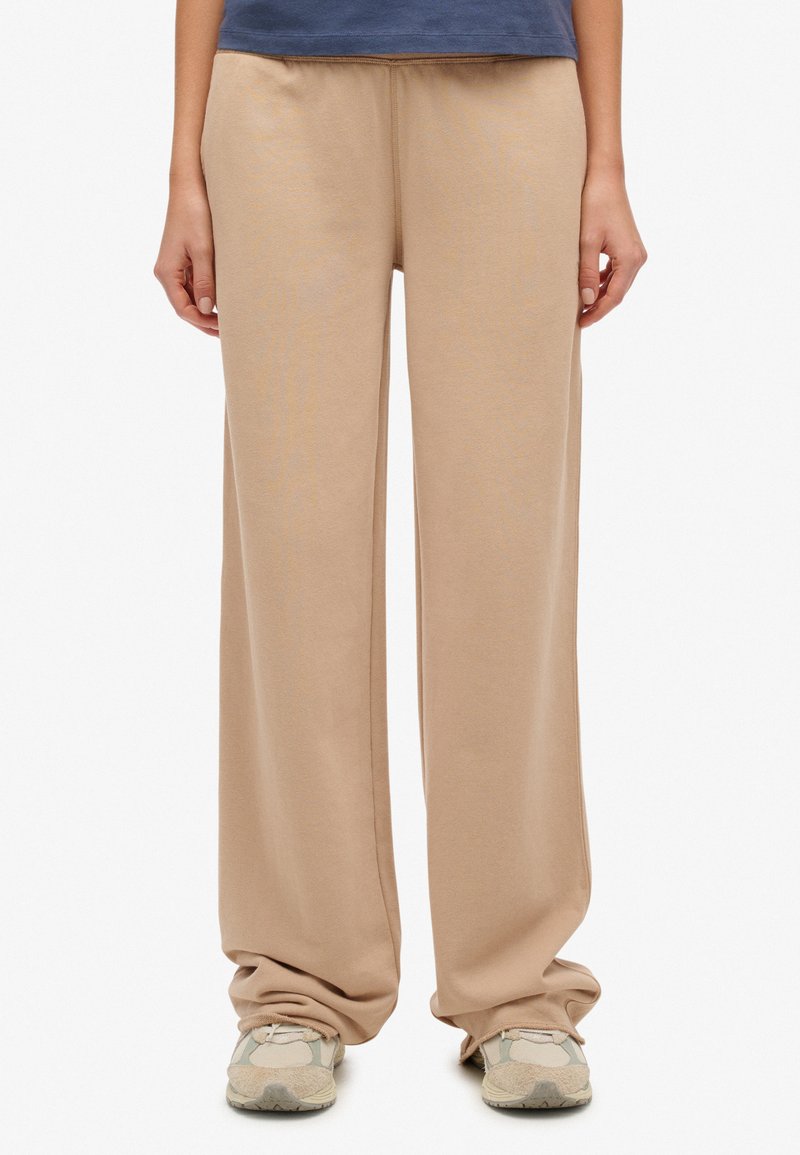 Beżowe szerokie spodnie dresowe o miękkiej fakturze i elastycznym pasku w talii, noszone ze szarymi tenisówkami. Gładki materiał bez widocznych wzorów ani zdobień.