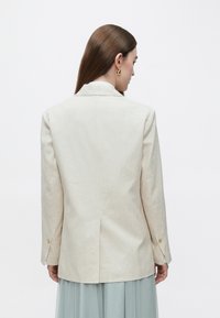 Beežist linase kangaga jakk notsitud kraega, ühe tagumise lõhikuga, varrukatel kuldsete nuppudega ja minimalistliku disainiga, kantud helesinise seeliku peal.