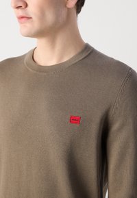 Brun strikket genser med rund hals og en rød rektangulær logo-patch på venstre side. Glatt tekstur med en tettsittende design.