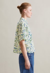 Kurzärmlige Bluse in Gelb mit grün-blauem Blumenmuster; verfügt über einen knöpfbaren Kragen und eine lockere Passform. Getragen mit Jeans.