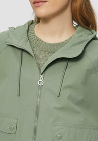 Veste zippée vert clair avec capuche, dotée de deux poches avant et de cordons de serrage. Portée par-dessus un pull en tricot vert texturé.