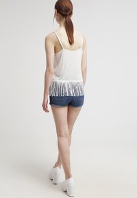 Biały top na ramiączkach z frędzlami na dole, zestawiony z dżinsowymi szortami. Tkanina jest lekka, a top ma cienkie ramiączka. Białe buty na platformie.