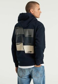 Felpa blu navy con una grande stampa grafica sul retro in rettangoli beige e grigi con testo nero. Realizzata in tessuto morbido, include un cappuccio.