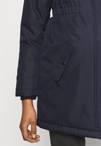 Laivastonsininen eristetty takki, jossa on vyötäröltä kavennettu malli, pitkät hihat ja sivutasku, jossa on musta painikenappi sulkemista varten.