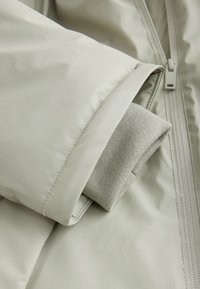 Prim-plan al mânecii unei jachete bej cu detaliu de manșetă tricotată și fermoar pe suprafața țesăturii.