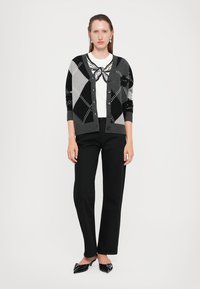 Sort og grå argyle mønster cardigan med knapper, båret over en hvit skjorte med grafisk design, kombinert med svarte bukser og sko.
