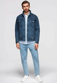 Giacca di jeans blu scuro con chiusura a bottoni e tasche anteriori; indossata sopra una camicia a righe blu chiaro. Jeans blu chiaro e sneakers bianche completano il look.