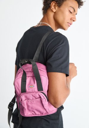 Jeune personne portant un t-shirt noir, avec un petit sac à dos rose Dickies sur une épaule, regardant vers le bas et légèrement de côté.