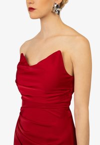 Rotes Satin-Kleid mit Herzausschnitt, das weiche Drapierungen an der Brust und eine taillierte Silhouette aufweist. Silberne, strukturierte Ohrringe setzen einen Kontrast.