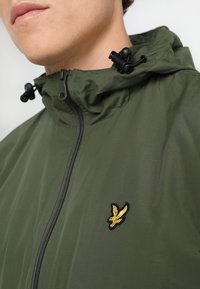 Veste à capuche verte avec fermeture éclair à l'avant, cordons ajustables noirs et petit emblème d'oiseau jaune sur la poitrine, portée par une personne.