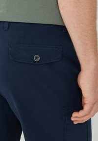 Bermuda cargo blu navy in misto cotone, dotate di una tasca posteriore con bottone e dettagli cuciti, mostrati da un angolo laterale con una mano appoggiata.