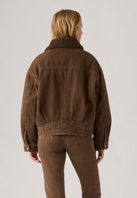 Persoon in een bruine jas met sherpa-kraag en bijpassende bruine broek, staand met de rug naar de camera tegen een lichte effen achtergrond.
