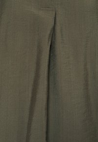 Pantaloni verde oliva con un tessuto liscio e strutturato. Presentano una leggera lucentezza e una parte frontale piatta con un dettaglio di tasca singola.