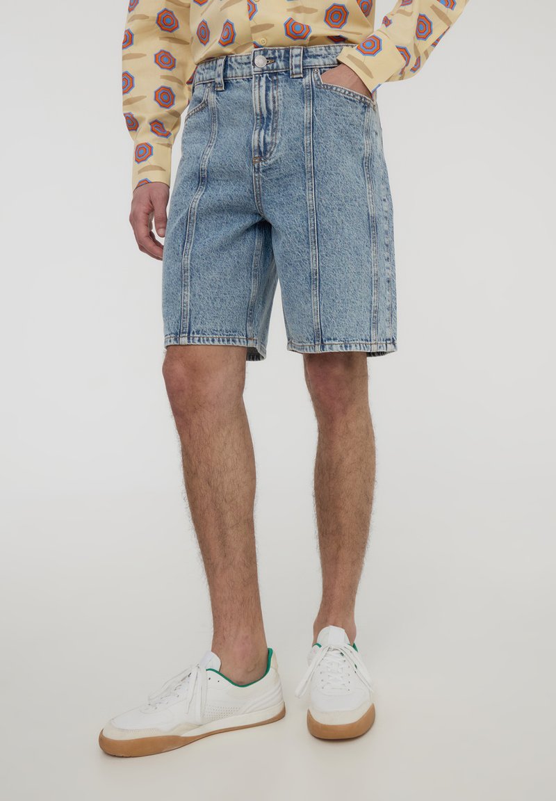 Shorts in denim azzurro chiaro con vita alta, con zone sbiadite e cuciture visibili. Indossati con sneakers bianche e una camicia a fantasia.