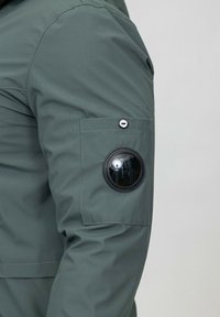 Veste verte à la texture lisse, dotée d'un patch noir rond sur la manche gauche et d'un bouton pression sur la poche au-dessus.