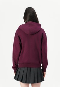 Person with long dark hair wears a purple hoodie and a gray pleated skirt, standing facing away against a plain white background.