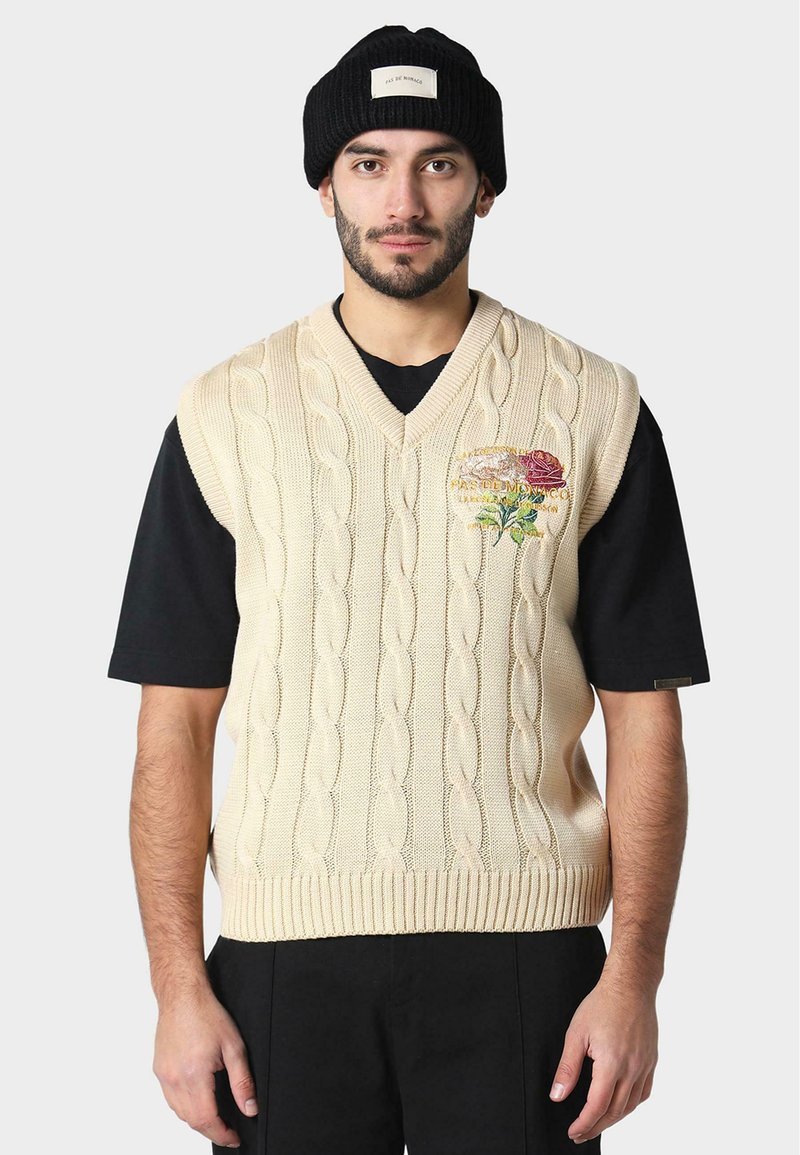 Béžový pletený svetr s kabelkovým vzorem a výstřihem do V; zdobený barevným květinovým vyšíváním na levém prsu. Nosit černou košili a čepici.
