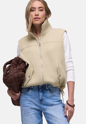 Donna che indossa un gilet senza maniche beige con zip sopra una camicia bianca a maniche lunghe, che tiene una grande borsa trapuntata marrone, abbinata a jeans azzurri.