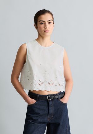 Femme aux cheveux bruns portant un crop top blanc sans manches avec un motif ajouré et un jean taille haute foncé, debout devant un fond gris uni.