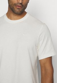 Camiseta de algodón de cuello redondo en color crema claro con mangas cortas. Presenta un pequeño logo de Nike en blanco en el pecho. Textura suave.