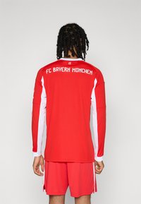 Sarkans FC Bayern München futbols krekliņš ar garām piedurknēm, ar baltiem akcentiem un komandas nosaukumu aizmugurē baltā burtnīcā.