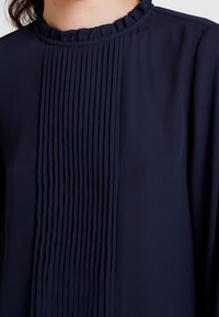 Blouse bleu marine avec devant plissé, tissu texturé et col à volants. La chute fluide et la coupe ajustée mettent en valeur le design.