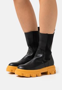 Bottes en cuir noires mi-mollet avec des semelles épaisses et chunky de couleur orange vif, portées sur des jambes nues devant un fond uni.