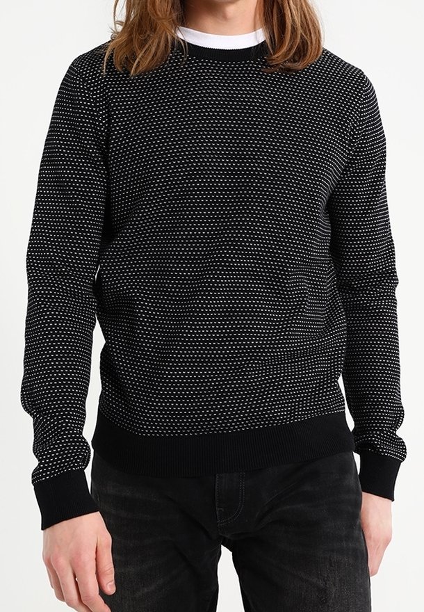 Homme portant un pull noir à petits pois blancs, assorti d'un pantalon noir, debout devant un fond blanc uni.