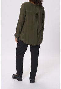 Femme aux cheveux bouclés portant une blouse à manches longues verte olive, un pantalon noir et des mocassins noirs, se tenant devant un fond blanc.
