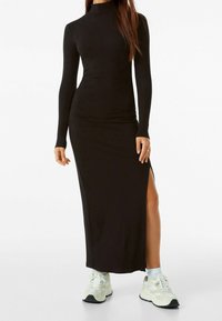 Robe maxi noire côtelée avec manches longues et col montant ; présente une fente latérale et est associée à des baskets blanches et grises.
