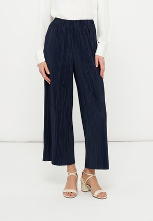 Femme portant un pantalon large plissé bleu marine associé à un haut blanc à manches longues et des sandales à talons blocs beige sur un fond uni.