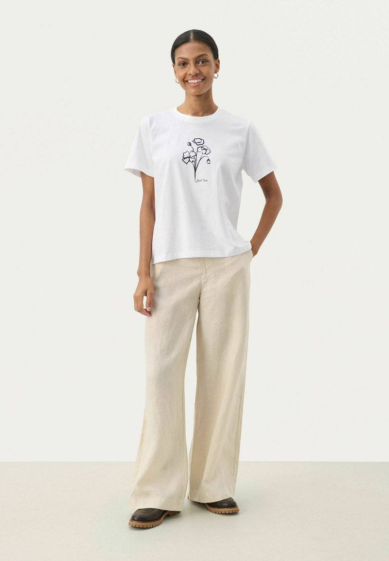 Weißes Baumwoll-T-Shirt mit einem schwarzen floralen Grafikdruck, kombiniert mit weiten beigen Leinenhosen und braunen Schuhen mit rutschfester Sohle.