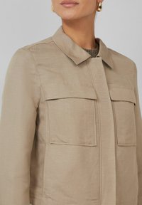 Beige lichtgewicht jas met een opstaande kraag, twee borstzakken en een gestructureerde afwerking. Rechte snit en minimale hardwaredetails.