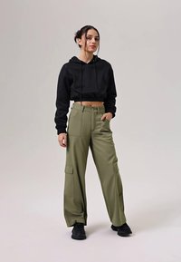Zwarte cropped hoodie met trekkoorden, gecombineerd met olive cargo broeken met wijde pijpen en voor- en zijzakken. Zwarte sneakers maken de look compleet.